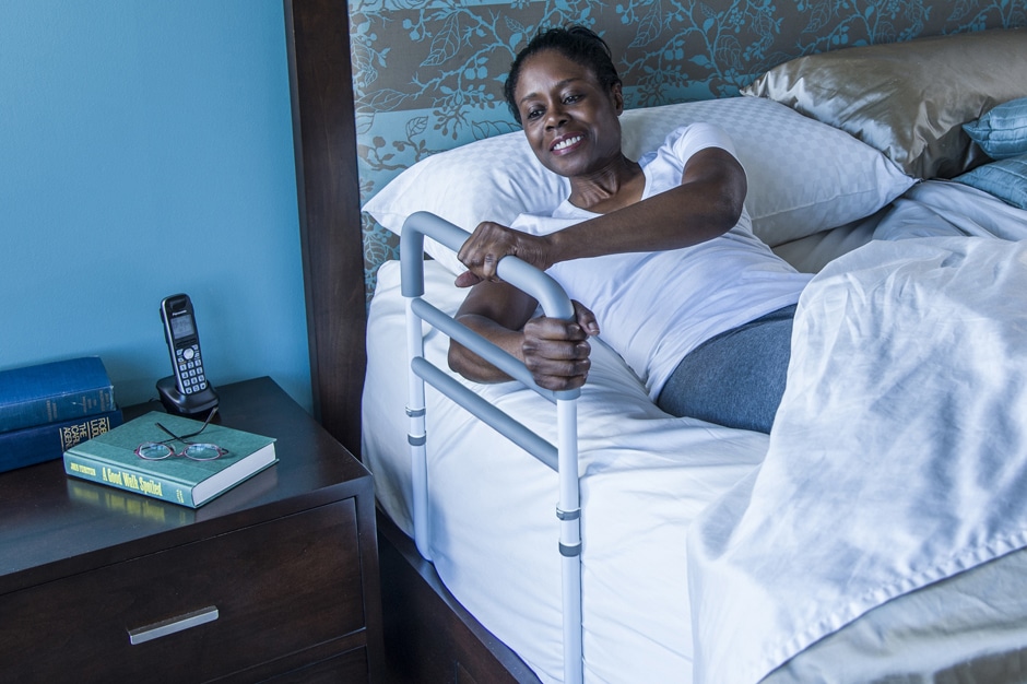 Photo of a woman in bed using a traditional bed rail (the HealthCraft Assista-Rail) to sit up in bed.