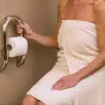 A woman holding Invisia wall toilet paper roll holde for support while sitting on a toilet.