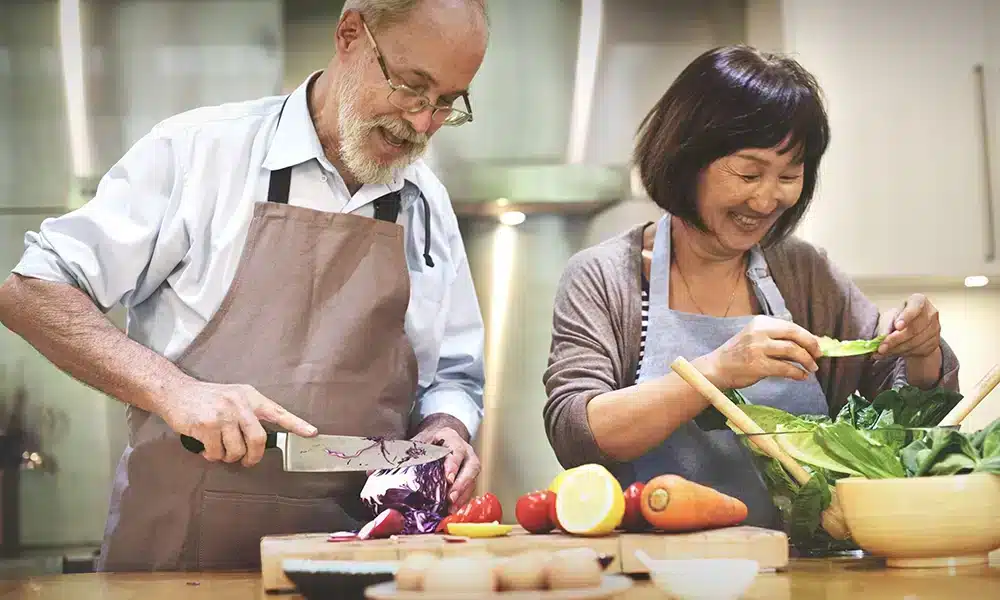 A senior couple preparing food together. A man is cutting cabbage while a woman is preparing lettuce.