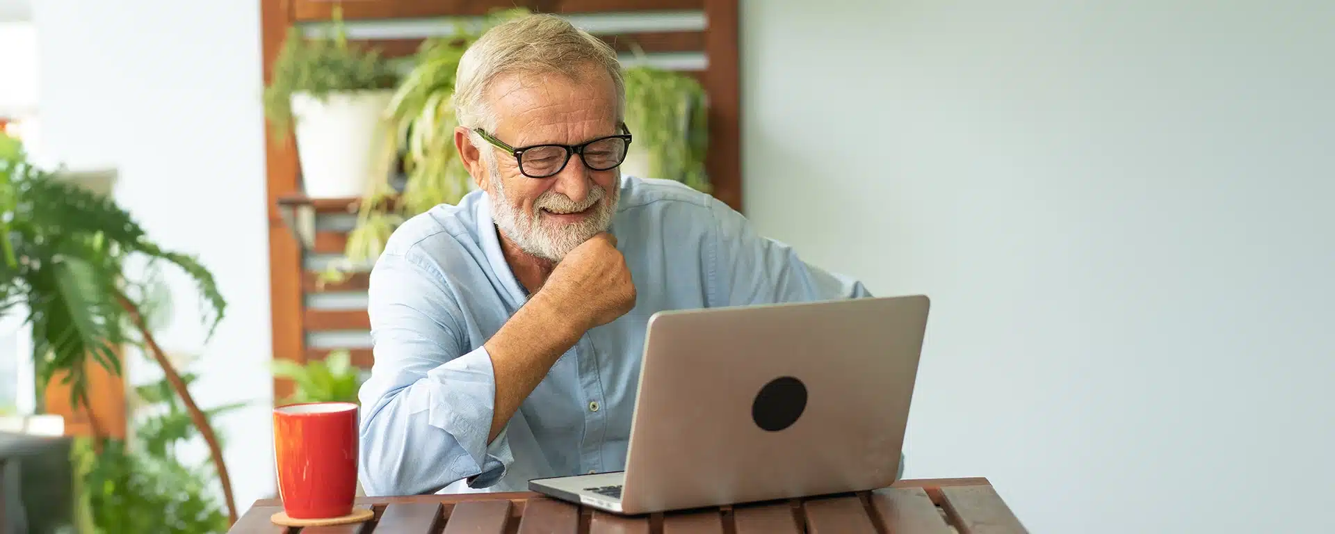 An older man smiling, having a coffee while using a laptop.